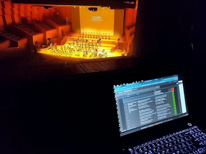 Cues on a laptop at Megaron, the orchestra and the surtitle screen beyond.