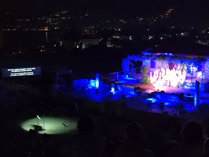 An open-air stage at night, titles on a screen beside the set.